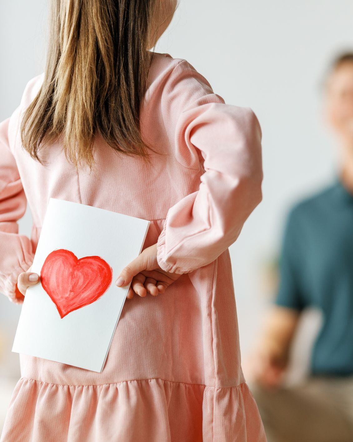 Little girl hiding a card decorated with a heart behind her back Little girl hiding a card decorated with a heart behind her back